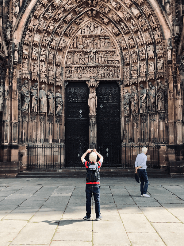 Joueur prenant une photo devant la porte sculptée de la Cathédrale de Strasbourg dans le cadre du jeu de piste "Les Gardiens de la Cathédrale", une aventure interactive de découverte du patrimoine.